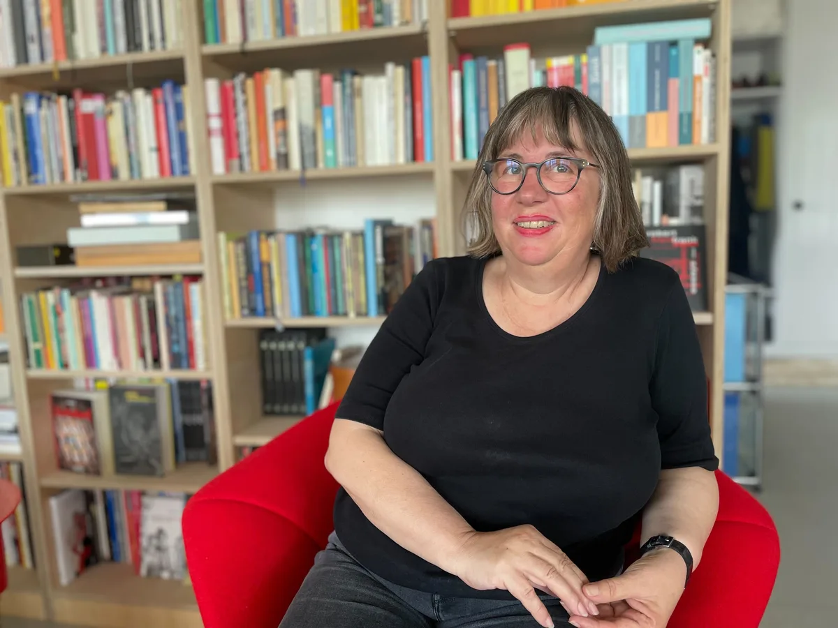 Judith Stofer hat eines der Hochbeete bepflanzt. Sie lebt seit drei Monaten hier.   Frau mit Brille sitzt in einem roten Sessel vor einem Büchergestell und schaut in die Kamera.